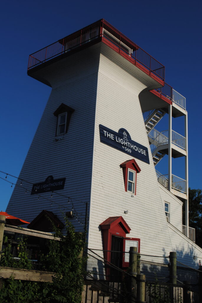 A large white lighthouse with red ornaments around the few windows and at the top. There's a blue sign "The Lighthouse by 540" and white stairs on the right hand side. On the bottom left side, there are lots of green plants and part of a red sunshade on a wooden patio.