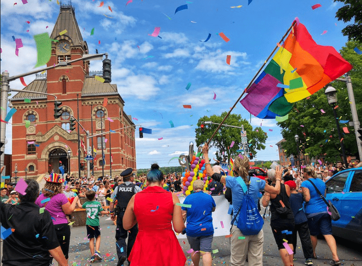 A crowd of people walking by City Hall, with lots of  large confetti pieces in the air. One person is holding a large rainbow flag.