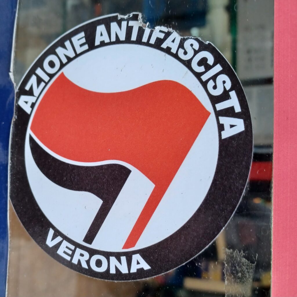 A black/white round sticker on a window that says "Azione antifascista" at the top and "Verona" at the bottom. There's a red/black flag in the middle as well.