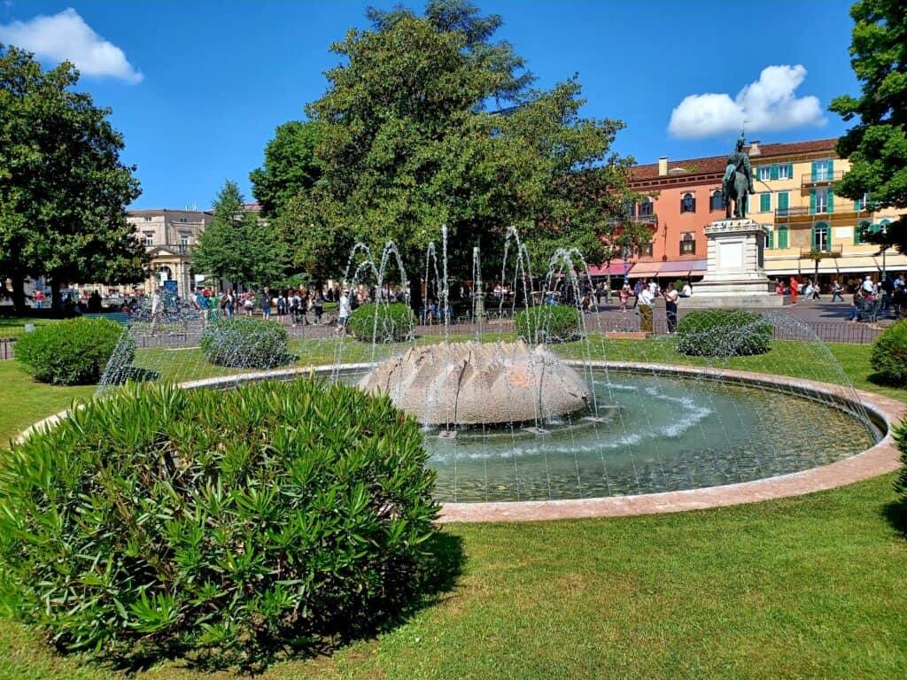 A nice green area with a bush in the foreground and green trees in the background. The fountain is in the middle and there's a statue and some colourful houses in the background as well.