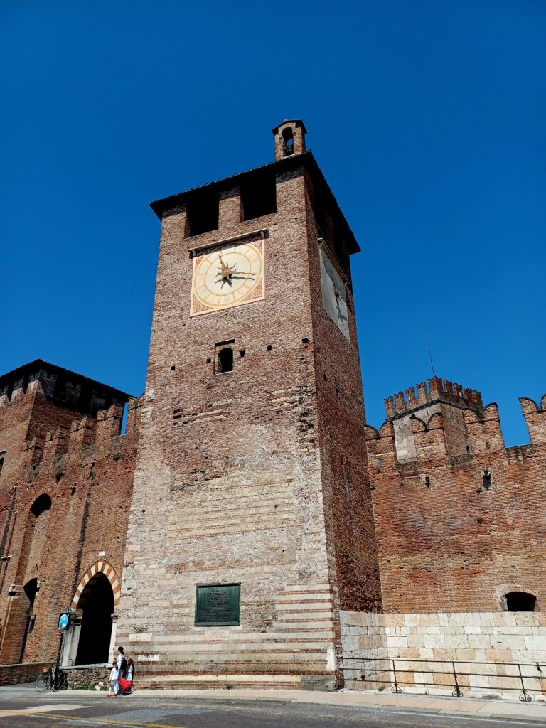 A high tower that's part of Castelvecchio. It consists out of mostly red brick stones and there's a gold/red brick coloured clock with with golden clock hands and very small tower at the very top. It's surrounded by a few m-shaped merlons and a few people are standing in front of the entrance to the castle complex.