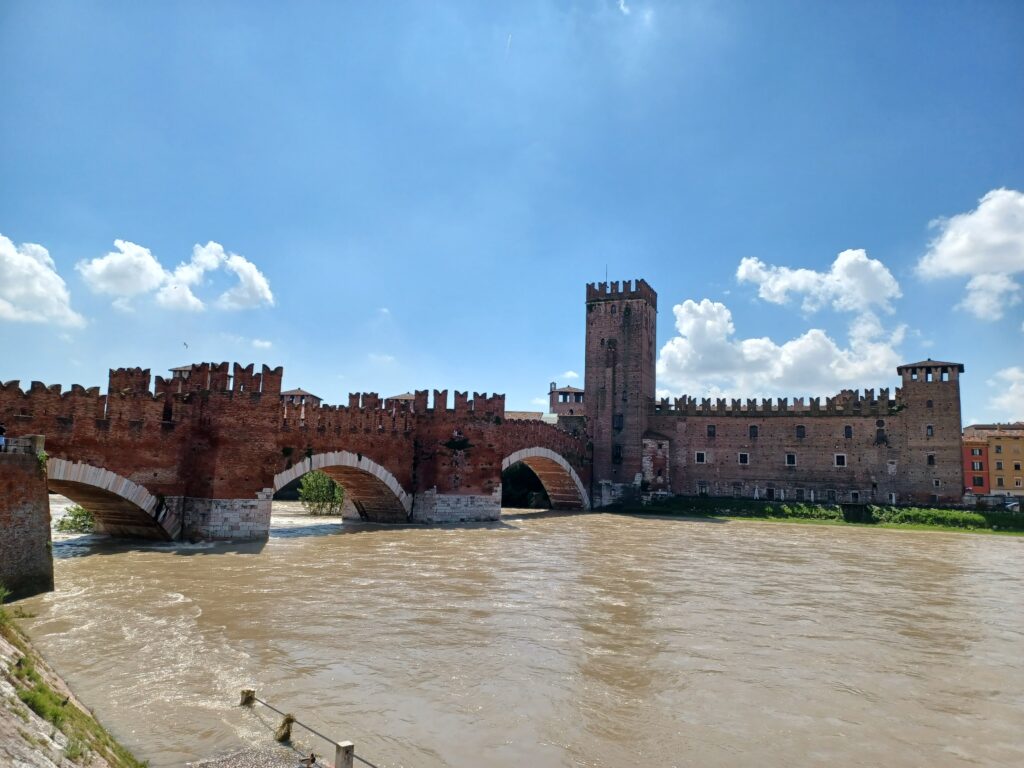 Adige River view of the Castelvecchio complex: The Scaligero Bridge with 3 arches is built out of red brick stones at the top, and white marble at the bottom. On the right, there's the Castelvecchio Castle with its largest tower on the left. The water is a bit high as well. 