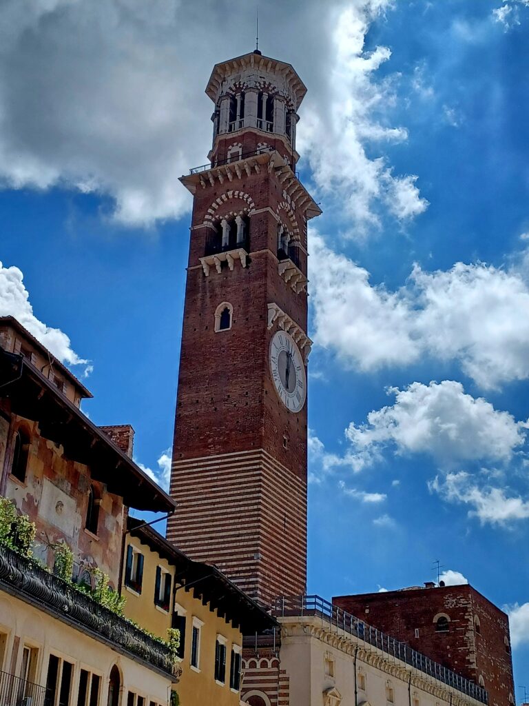 A skinny high bell tower with a large round white clock towards the bottom. It's mostly made out of red brick stones, but there's some white decor spread over the tower as well. Underneath the tower, there are some bright yellow houses and a one pink house with frescoes on it. 