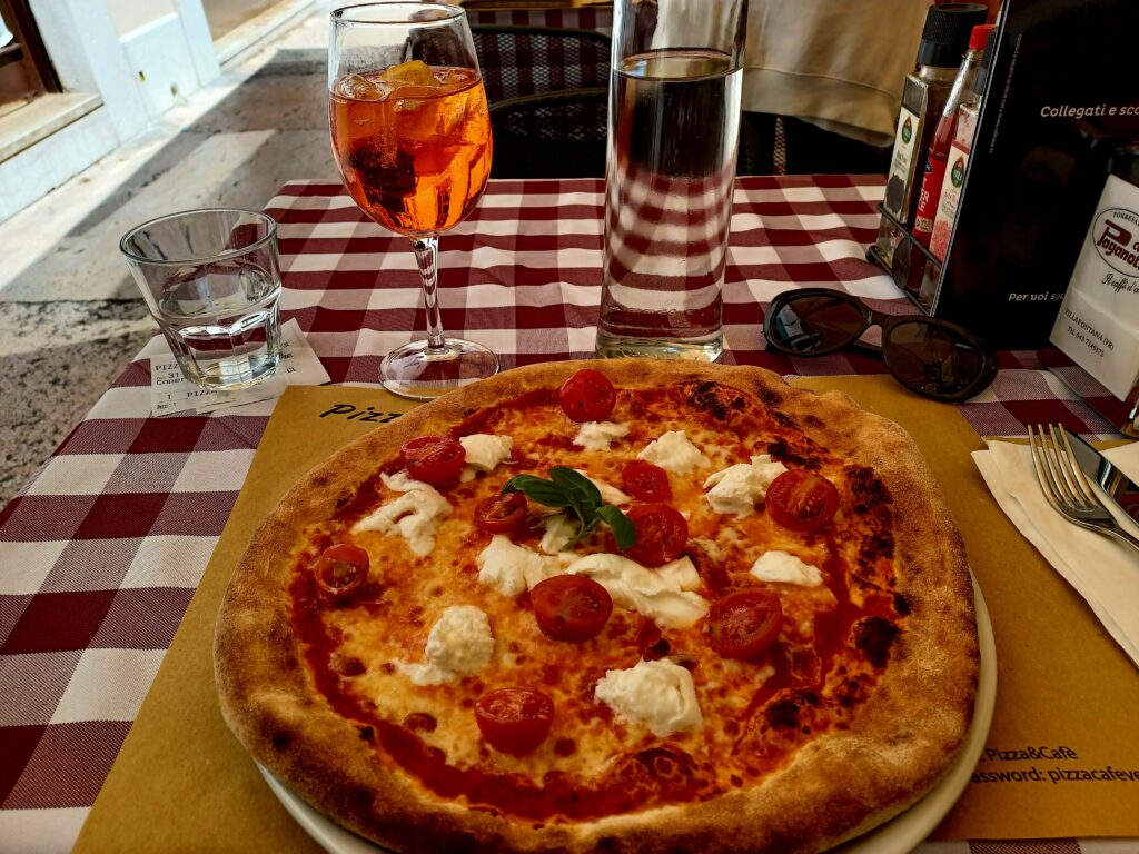 A large Italian-style pizza with cherry tomatoes, mozzarella cheese, cheese, and tomato sauce with a few basil leaves in the middle. At 11 o'clock, there's a small glass of water, and an an orange aparol spritz wine glass and a glass water bottle at 12 o'clock.