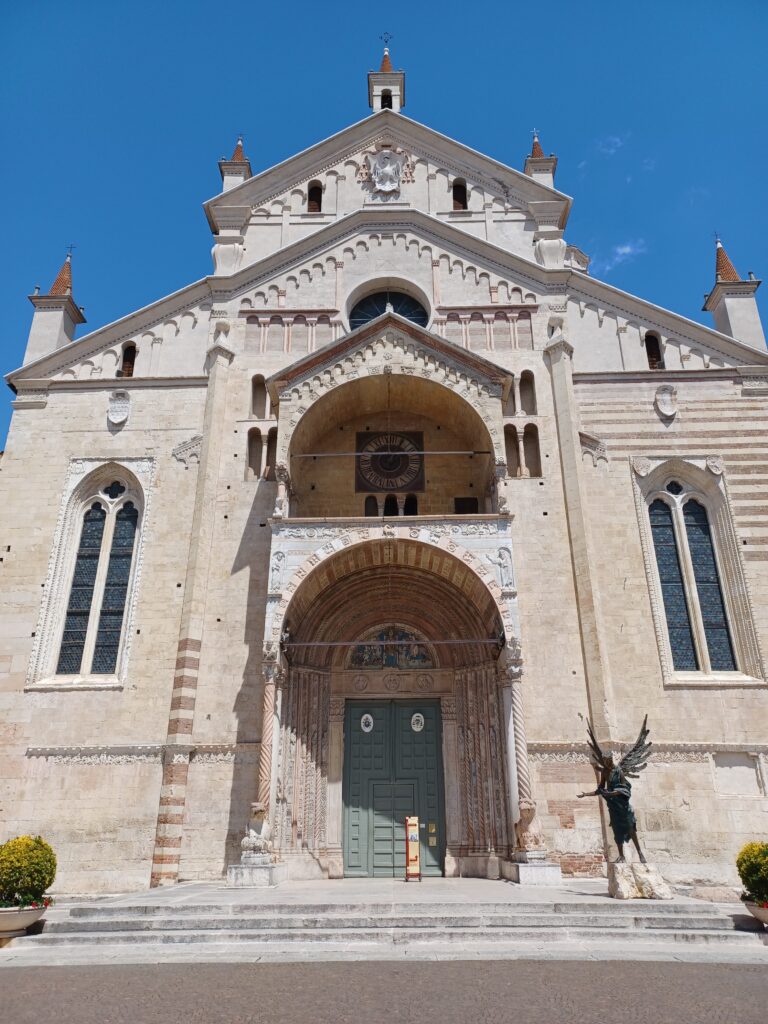 A high beige church building with 5 little towers (1 in the middle, and 2 towers on the left and right), a clock, high dark windows, and a dark green entrance door. There's a black sculpture with wings on the righthand side next to the entrance, and 3 sets of steps, and a green bush on both sides. 