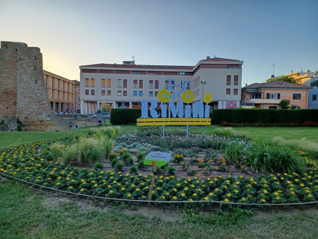 A light blue and yellow sign that says "Rimini" with a bike on top of it. There's a circular yellow flower bed around the sign as well. 