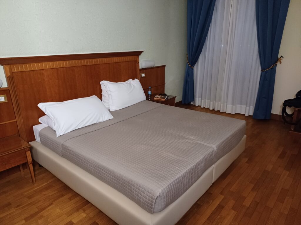 A queen size bed with 2 white pillows and a grey blanket. There's a brown wall behind the bed and blue and white blinds by the window next to the bed.