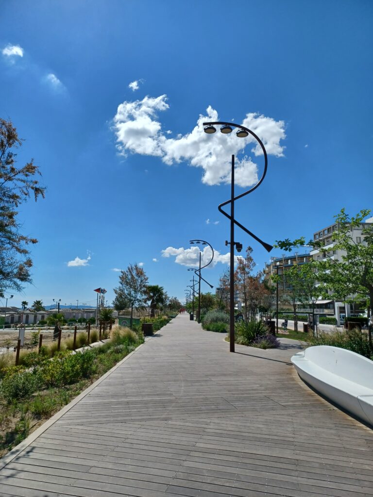 A long wooden pathway with black streetlights and trees/bushes on its left and right side.