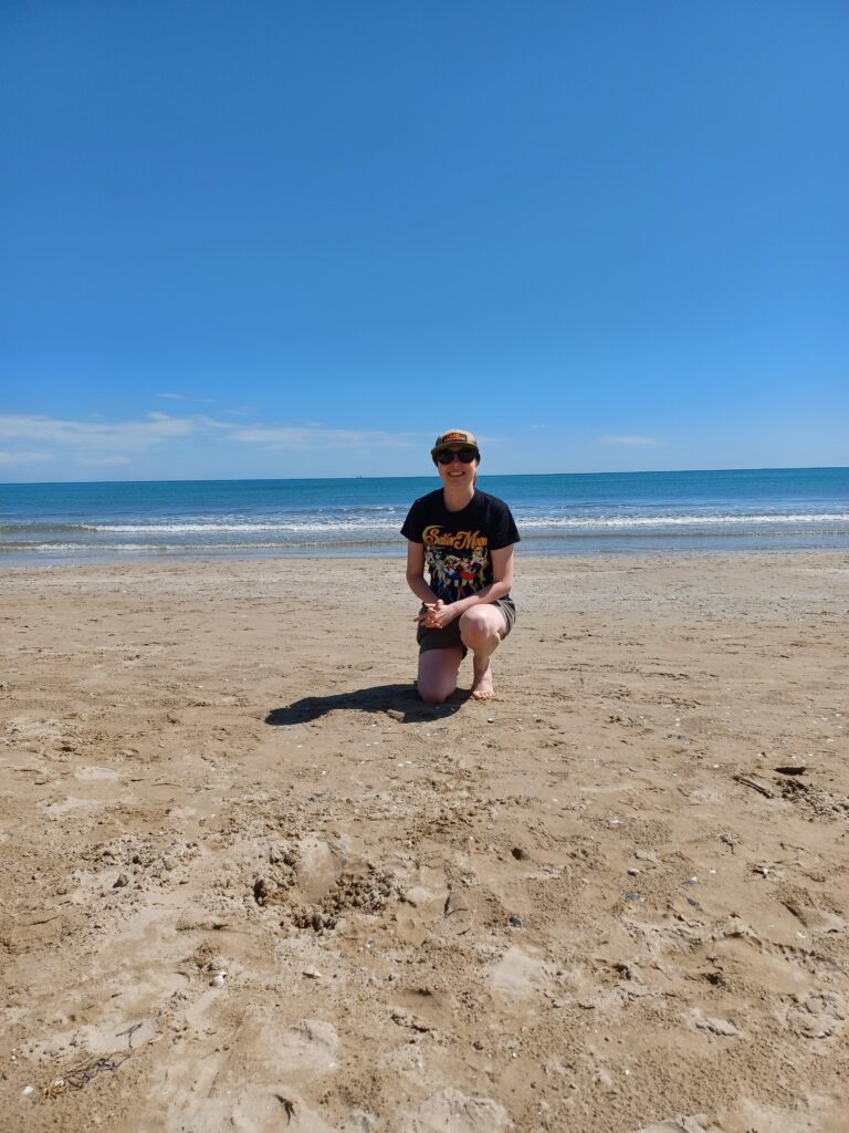 Me kneeling on the beach with the blue water of the Adriatic coast in the background. I'm wearing a green baseball cap, sunglasses, a black Sailor moon shirt, and beige shorts.