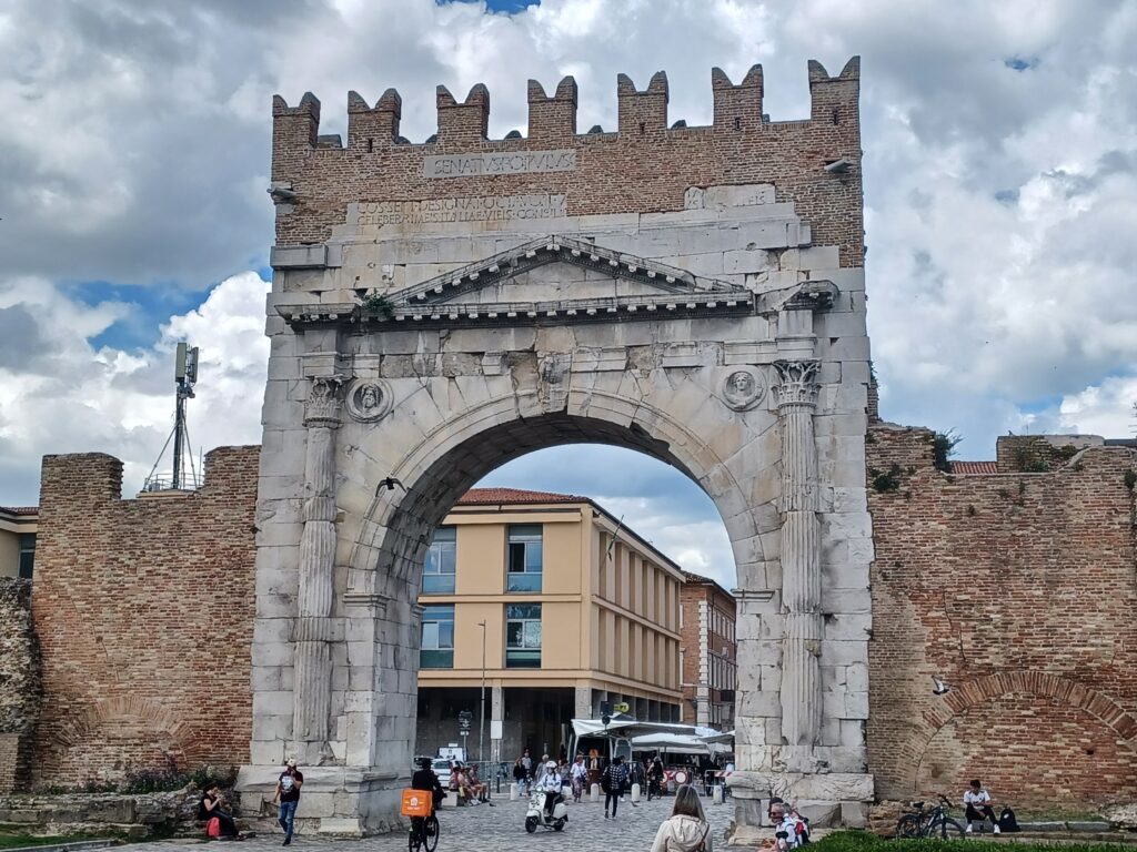 A high grey arch with red brick stone walls on its left and right side. There's a column on either side of the arch and people are walking/riding through the large gap. Some are sitting or standing on the right and left side as well.