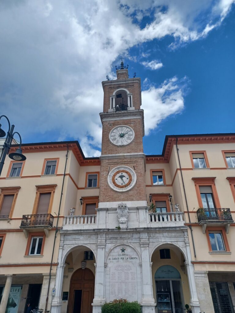 A skinny clock tower next to (what looks like) residential buildings. It has two clocks underneath each other, and is built out of brick stones. At the bottom, there are two arches on the left and right side, with a facade that says "Gloria ai Caduti per la liberta" and a bunch of names on it.