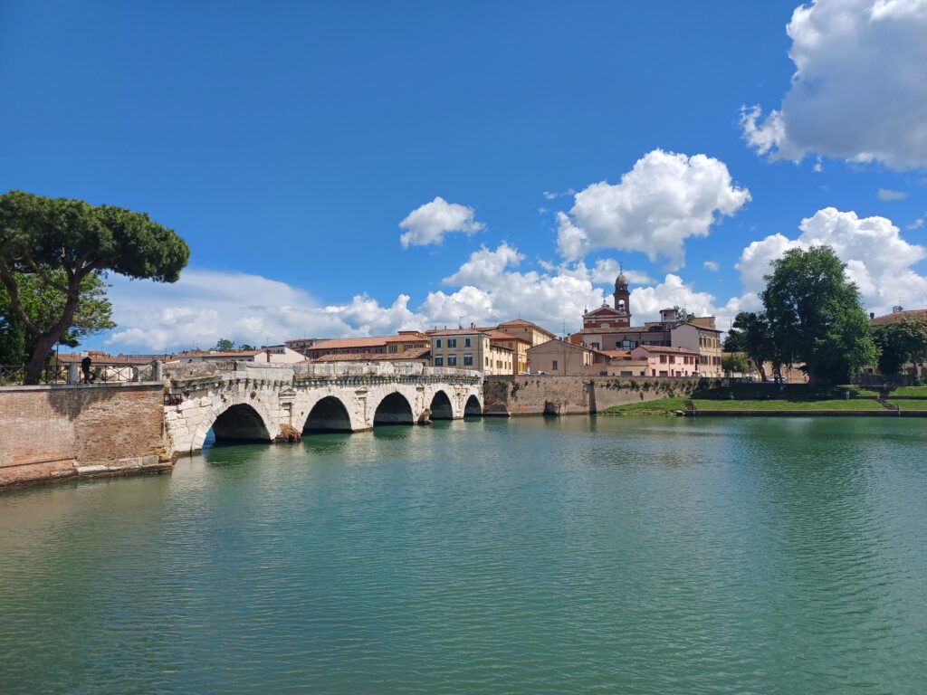 A beige symmetrical ancient bridge with 5 arches is crossing a green/blue river, with trees on the side of it. There are also a few houses and a high tower visible in the distance of the right hand side of the picture.