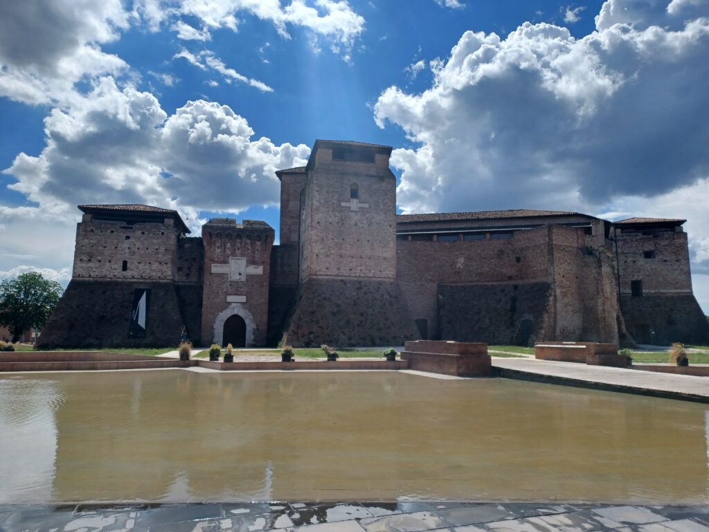 A castle with a 4 tower areas that all look different. The 3rd one from the left is the highest, and there's a water area, paved walkway, and a few green spots in front of the castle as well. 