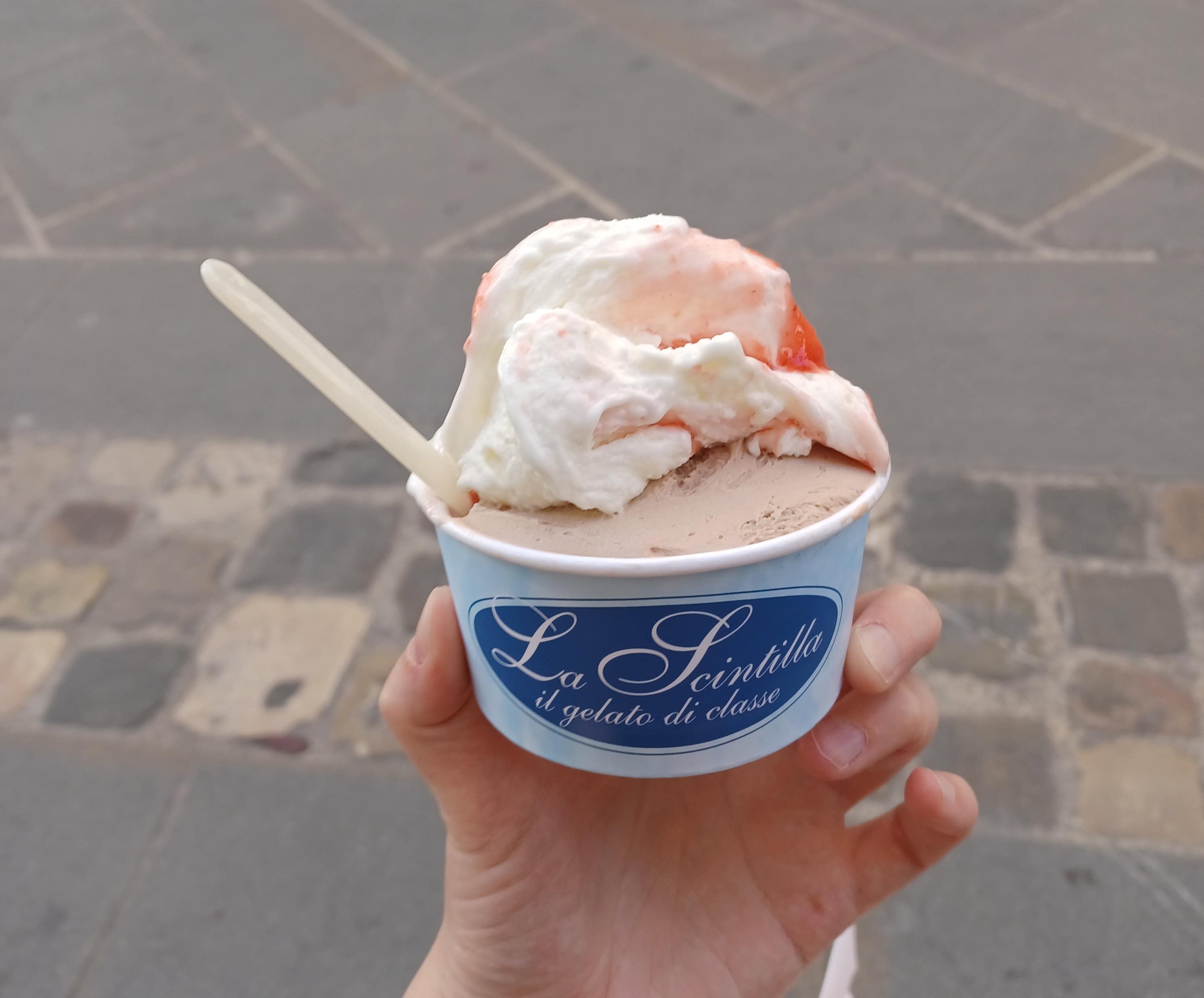 A white/blue cardboard cup of light brown (nutella) and white/red (cheesecake) gelato with a wooden spoon in my hand.