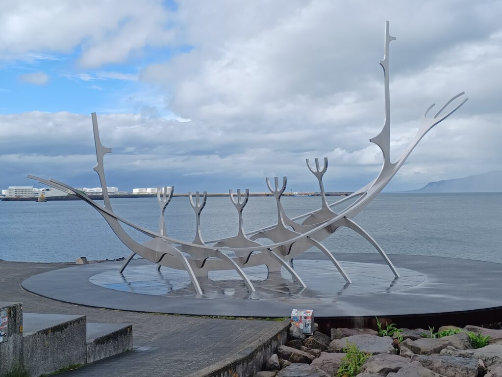 A white/grey dream boat sculpture in front of the sea. It stands on what looks like paddles, and there are several "forks" leading up to the sky.