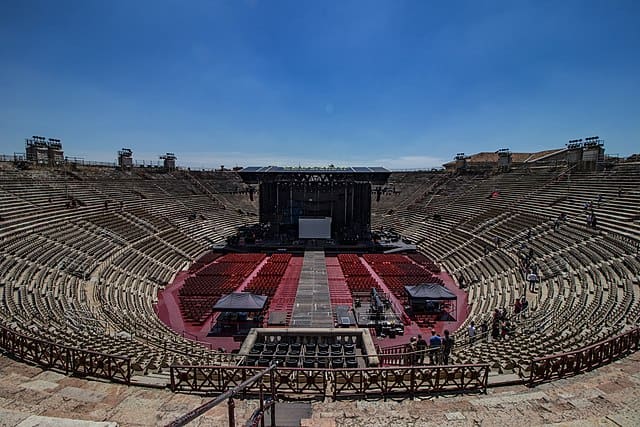 A circular arena from the inside, with a big black stage at 12 o'clock and many seats around it. There are some red carpeted sections in  front of the stage and two tented areas. A blue sky is in the background.