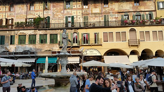 A busy market square with many people in the foreground. There are booths with white sunshades above them, and the grey Madonna Verona statue and fountain towards the middle. In the background, there's a yellow house wall at the bottom with green window shutters and a pink house wall with frescoes and green window shutters and some flowers and plants in front of it.