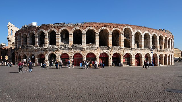 A circular ancient Roman arena from the outside with two floors, and lots of arches on each one. There are several people walking around in front of the arena. The building is mostly pink at the top and the rest is  a beige colour with a blue sky in the background.