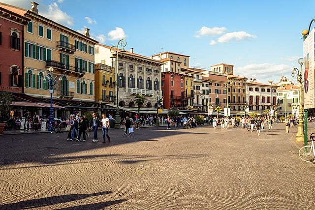 A buzzing square with people walking around on the left and right. There are some colourful buildings (red, orange, beige, yellow, and pink) mostly on the left side and little outside cafes and restaurants on the left with sunshades above them.