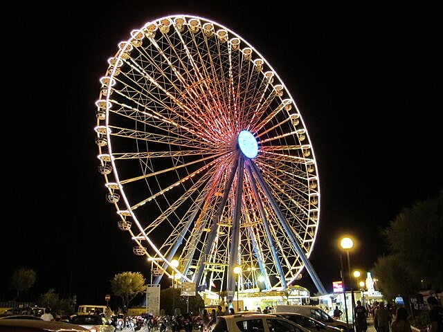 A large lit up ferris wheel is the focus of a picture taken at nighttime.