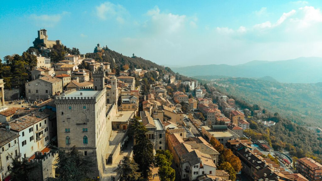 A drone view of San Marino with brown/grey castles, houses, and lots of trees, forest and some hilly areas.