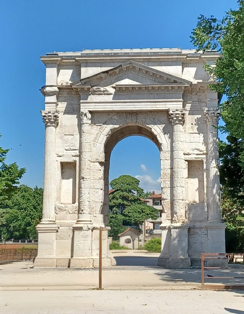 A large white Roman arch sits in front of a green area and a tree is on its right. There are two columns on each side and a few houses in the background.