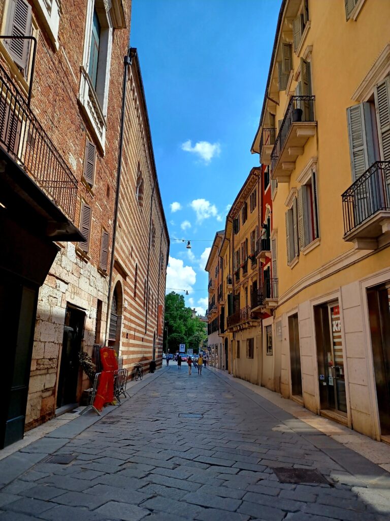 A cobblestone street with a few people walking in the distance. On the right, there are yellow houses next to each other with blue window shutters and balconies. On the left, there's a red brick stone building, one has white stripes as well. 