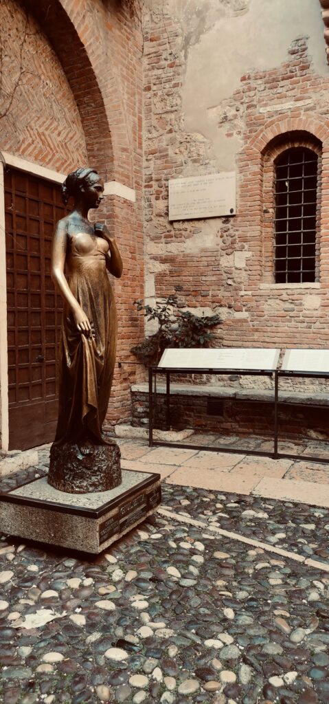 A large statue of Juliet, a teenage girl. She wears a strapless long golden dress and holds on to its bottom with her right hand. The left hand is touching her shoulder. Her hair is put up in a ponytail and she wears a hairband as well. 