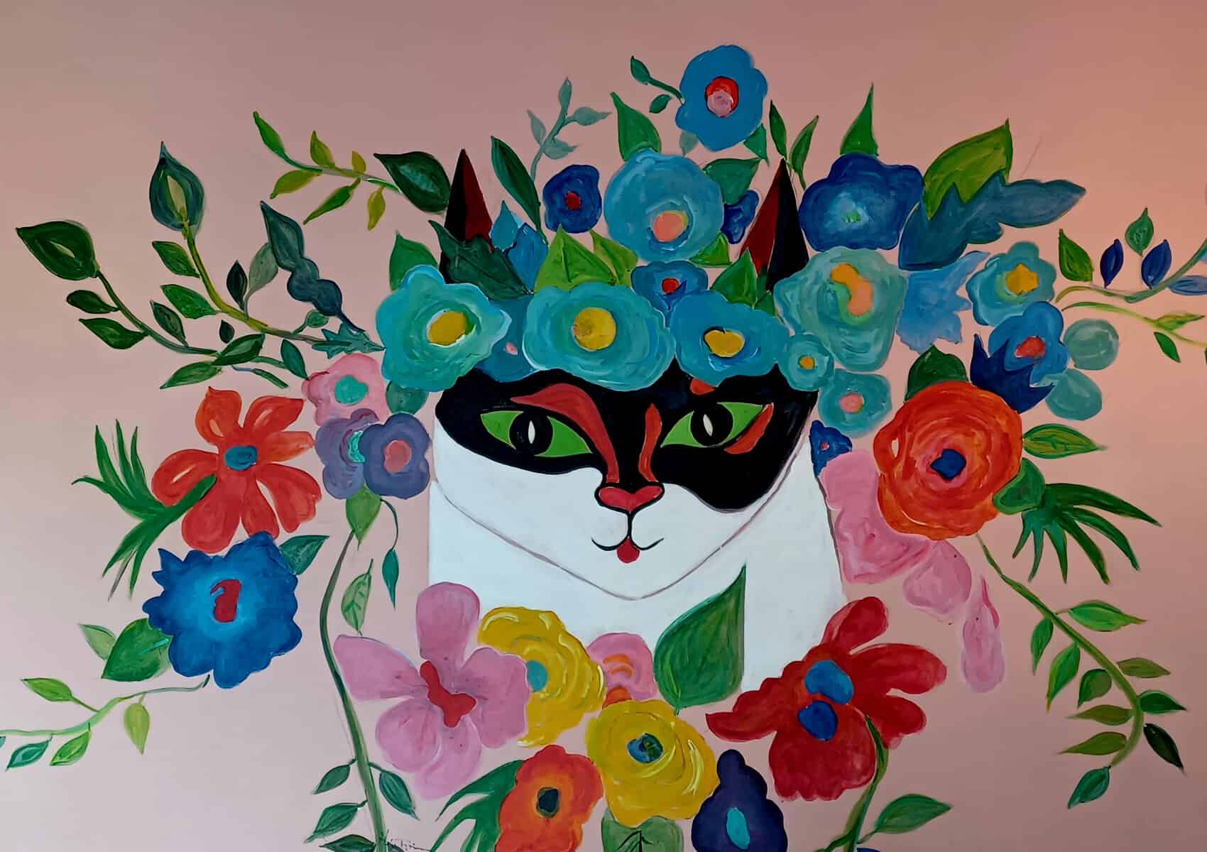 A painting of a white/black/red cat with green eyes on a rose wall. It's surrounded by flowers of various colours. It looks like it's wearing a flower crown as well.