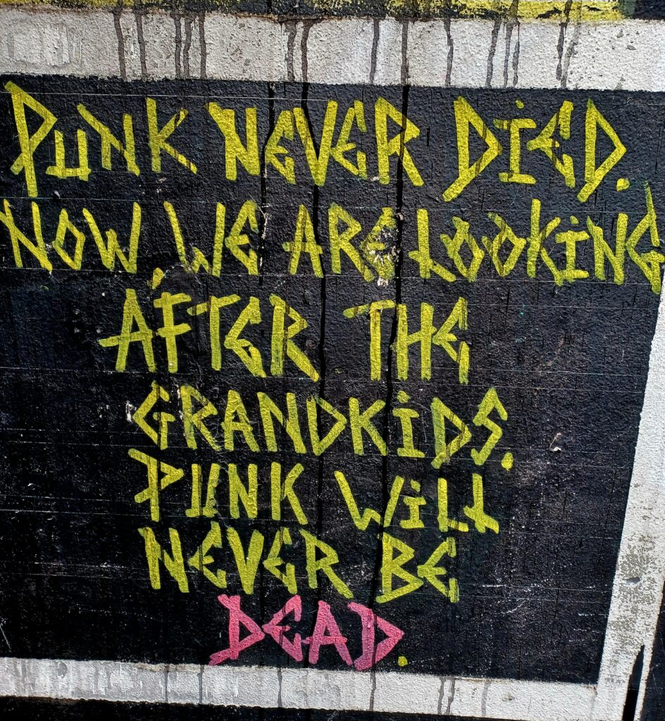 Just one graffiti sentence on a black wall, all written in uppercase yellow letters, except for the last word, which is pink. It says "Punk never died. Now, we are looking after the grandkids. Punk will never be dead."