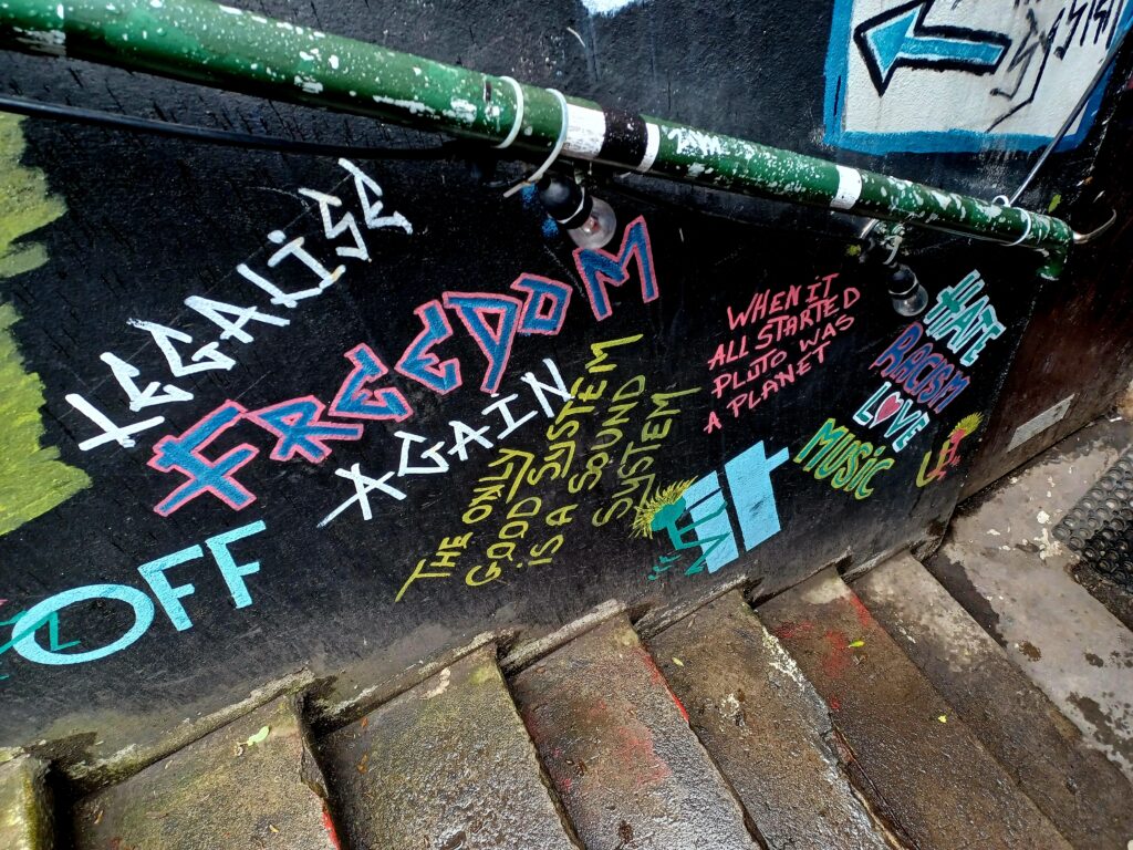 More colourful uppercase Punk graffitis on a black wall. A few example are "Freedom", "Again", "Hate Racism love music", and "When it all started Pluto was a planet". The colours are pink, white, yellow, blue, and green.