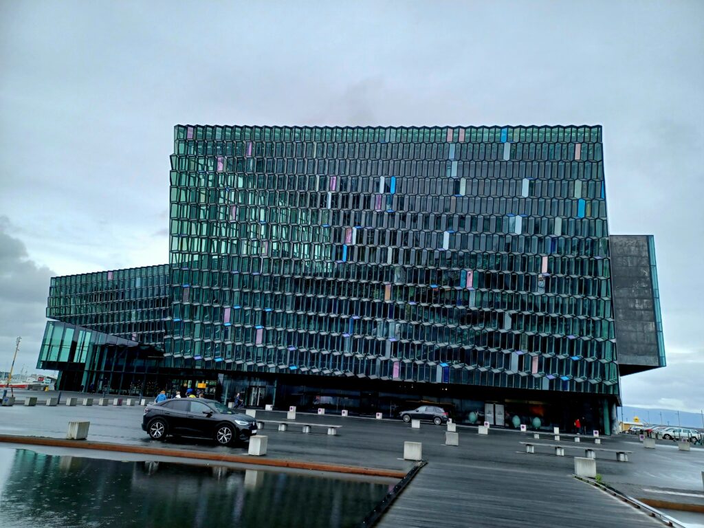 A large glass building with a honeycomb pattern. There are lots of these on the buildings, probably around 50! There are a few cars parked in front of the building. 