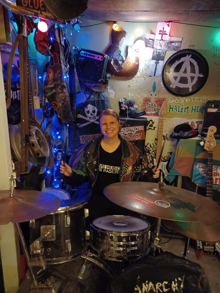 Me sitting behind the drums at the Icelandic Punk Museum. I'm wearing a black/red leather jacket with studs on it from the museum. There's a black/white anarchy sign, photos, a black pirate flag, and amps next to the wall. There's also a guitar in front of one of the amps behind the drums.