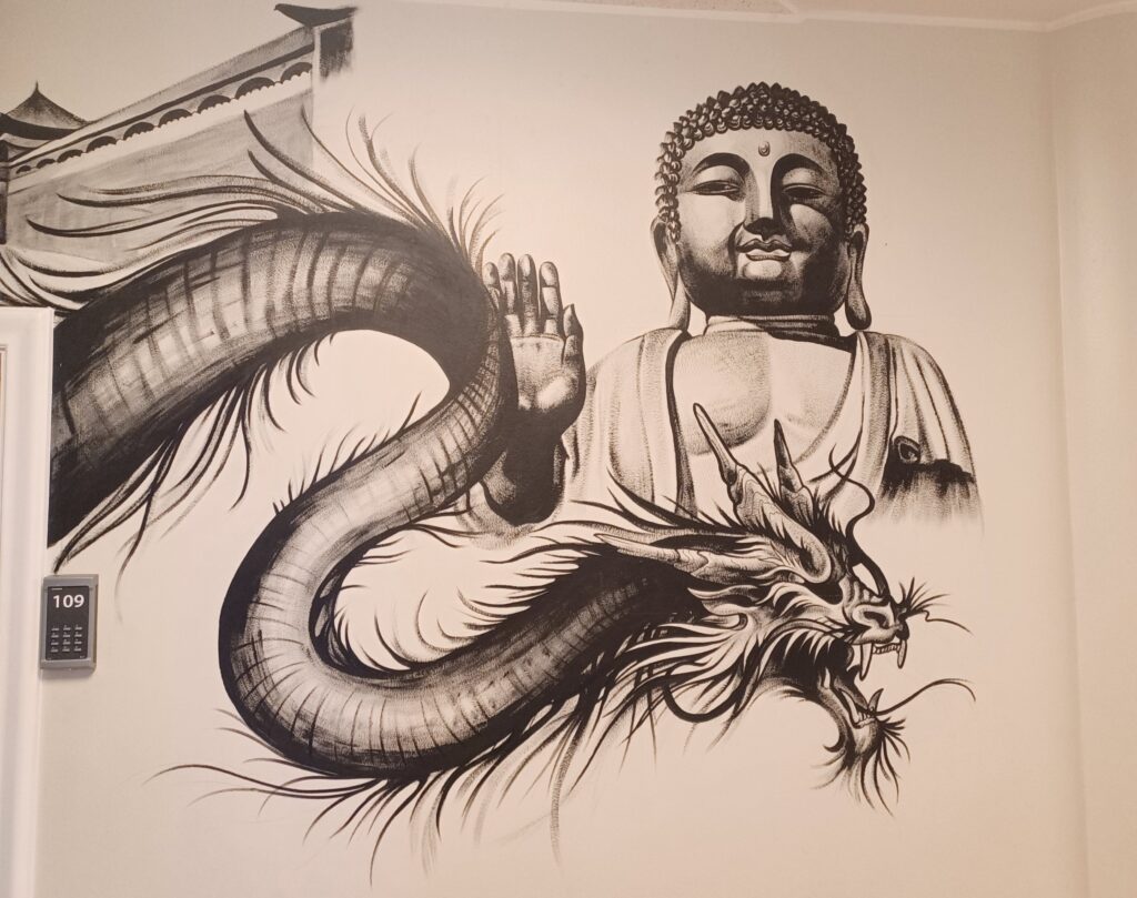 Black drawings of a Buddha statue and a large dragon on a white wall. In the background, there's a temple building (or something similar).