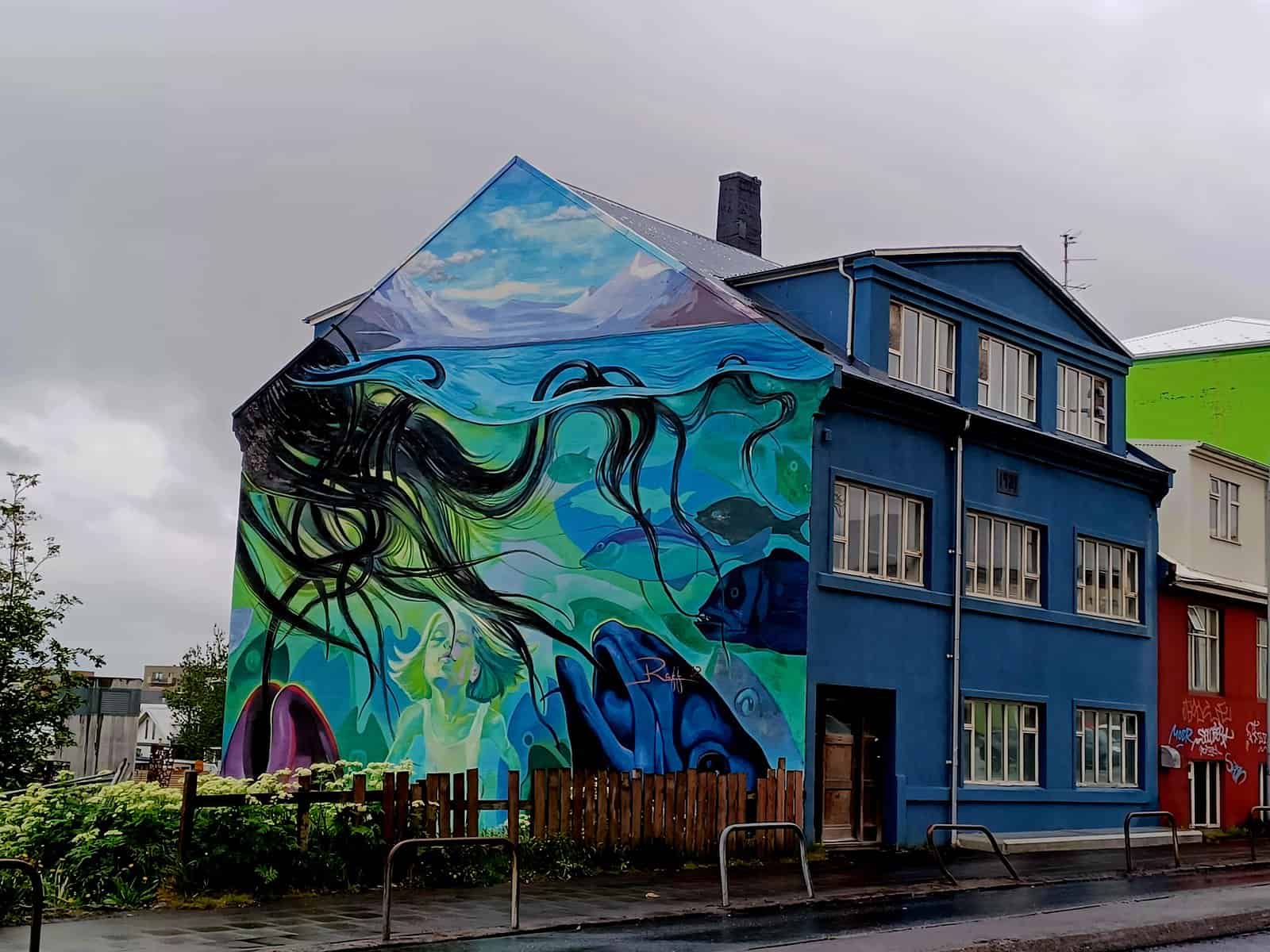 A bright blue house that has a mural on its left wall. Several dark blue fish are looking at a black octopus on the left side. Its tentacles look like seaweed or even black hair!