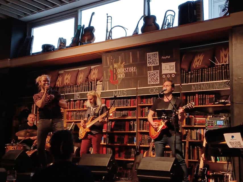 The Bookstore band performing. There's a singer, bassist, and guitarist in the front, and the drummer in the left back corner. There are several full book shelves on the wall behind the band. At the top of the shelf in the middle, it shows the band's name and logo.