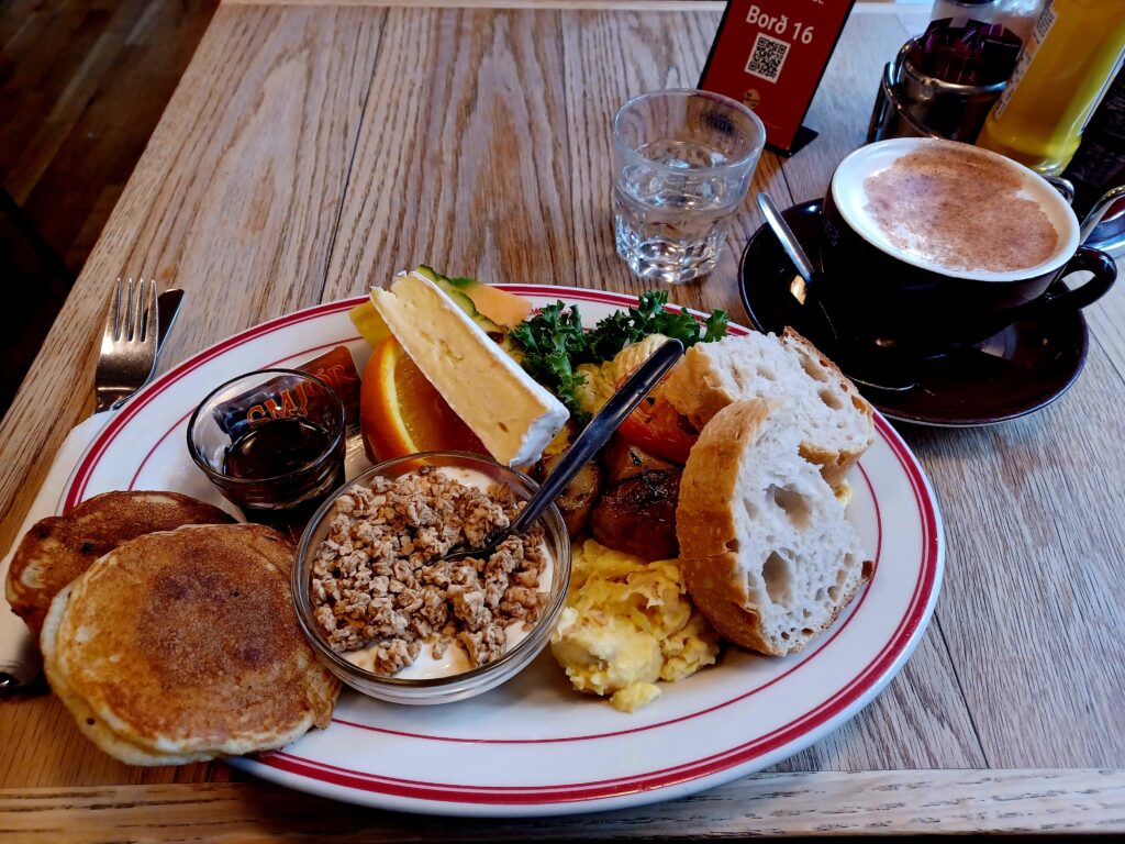 A large oval plate with brunch foods, e.g. scrambled egg, bread, cereal, yoghurt, pancakes, syrup, orange, and cheese. 