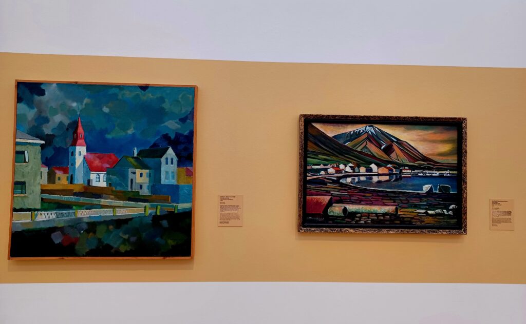 Two paintings on a yellow/white wall with city buildings (a tower and houses and a road) and a mountain and a lake. 