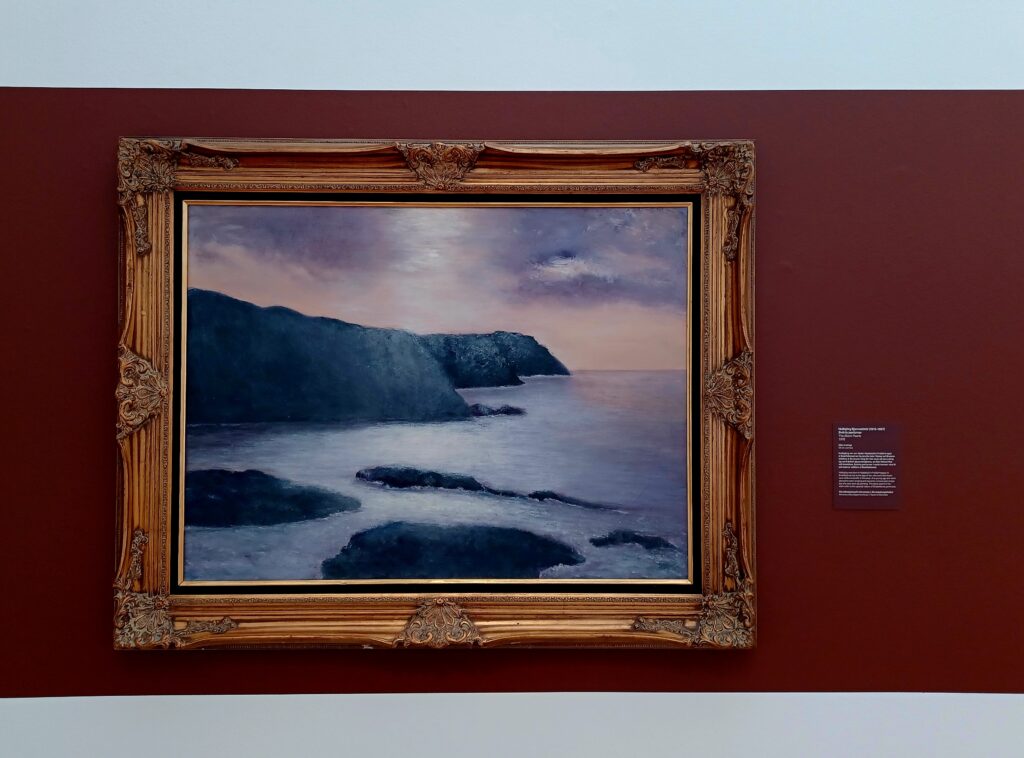 A framed painting on a red/white wall of a cliff and islands inside the ocean.