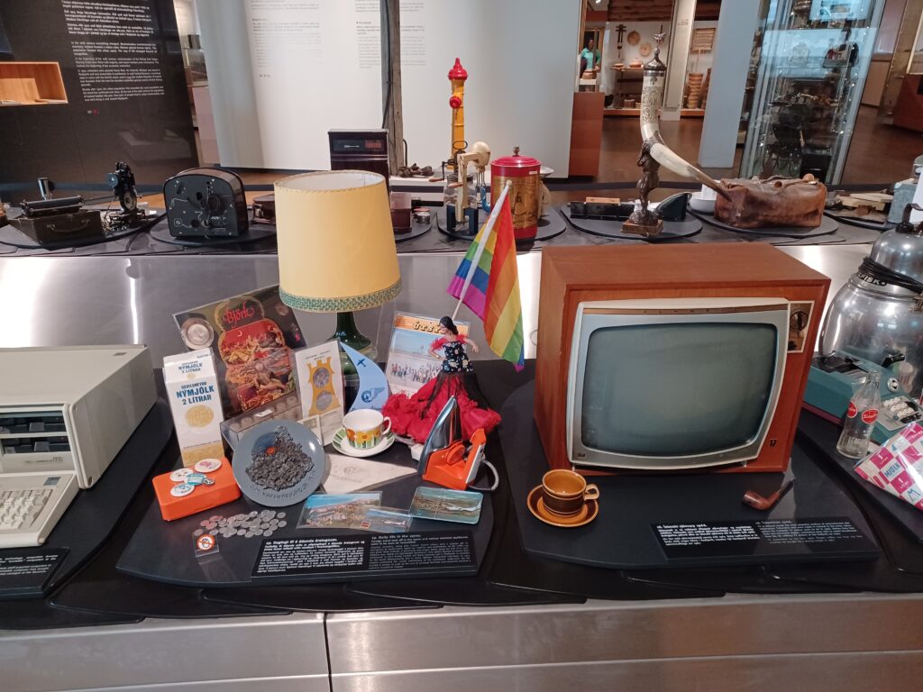 A display of items from the late 20th century, e.g. a small brown/orange tube TV, small mug with saucer, rainbow flag, a yellow nightstand lamp, grey ashtray, and half a printer on the left. 