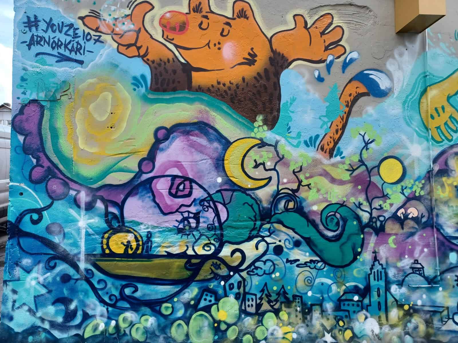 A colourful graffiti mural with an orange/brown pig?! You also see a crescent moon, and high tower, a boat, and some city skyscrapers. The colours are purple, yellow, light and dark blue, green/brown, and a bit of grey.