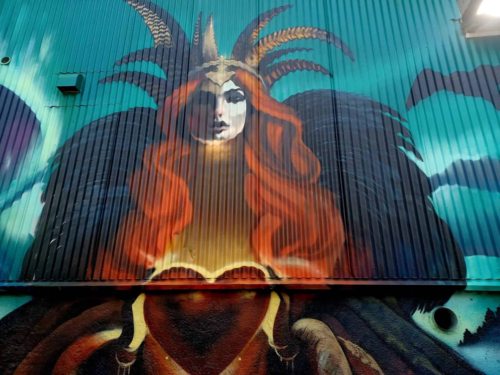 A painting of a red/orange haired woman that has several horns on her head, and black wings. It looks like she's holding a big red heart in her bands. There's a blue background as well.