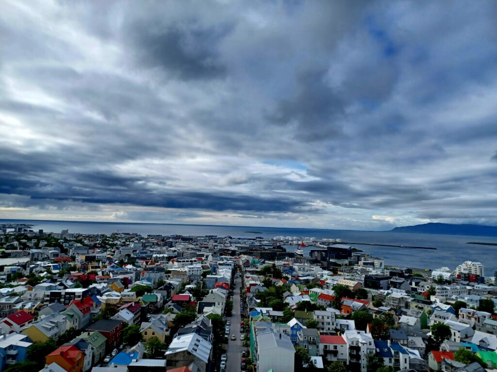 A bird's eye view of Reykjavik with many (colourful) houses, and cars driving on a large street. In the distance, there's a bit of blue sky, and the sea with some islands and a mountain on the right of the photo.