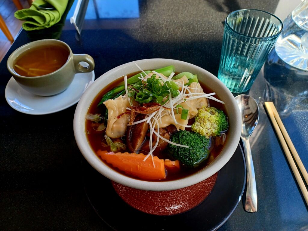 A wonton soup with broccoli, carrot, green onions, wontons, and noodles at the bottom. There's a ginger tea on the left and a glass of water on the right of the bowl.