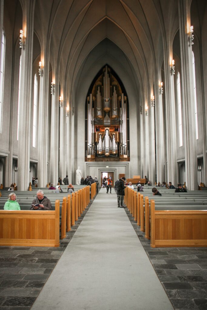 The inside of Hallgrim's Church. There are several rows of wooden benches on the left and right side and people are sitting on them. The middle pathway leads to an impressive group of organs. The walls of the church are white with a lamp at the top of each column. There are windows as well. 