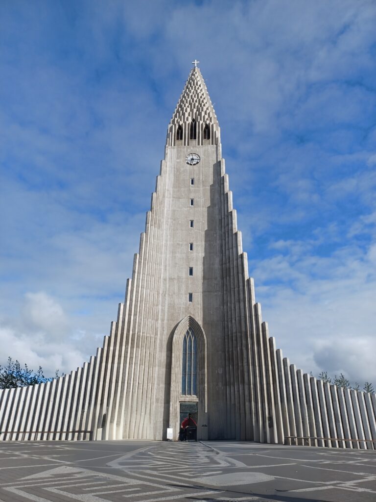 Hallgrim's Church on a sunny days with blue skies. It's grey and there are "steps" leading up to the top, as described above. There's a clock underneath the top and a white cross at the very top of the church. The entrance door is at the bottom and large window is above it.