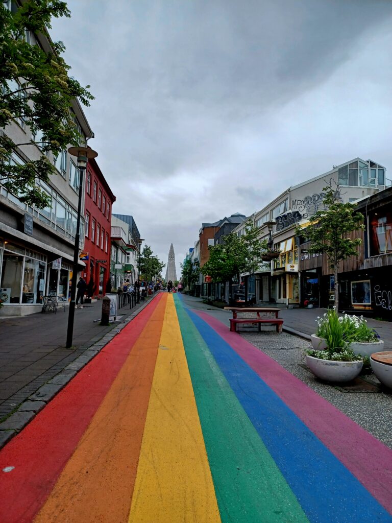 A long street painted in rainbow colours leading to Hallgrim's Church. There are a few plants/flowers, trees, picnic table, bike stands, and street lantern on the left and right side. Also lots of stores and a few colourful houses! People are walking in the distance, too.